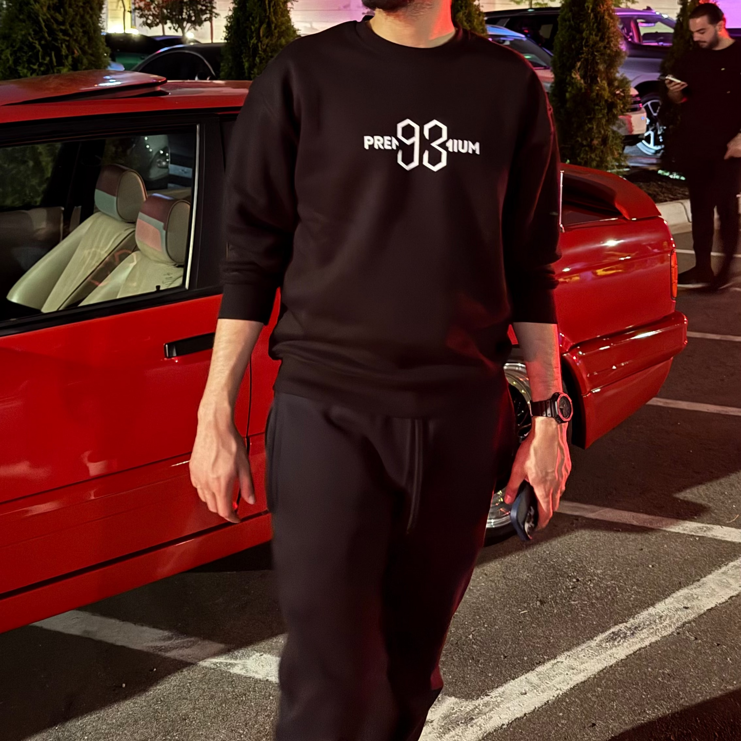 Man in a black sweatshirt standing next to a red car at night.