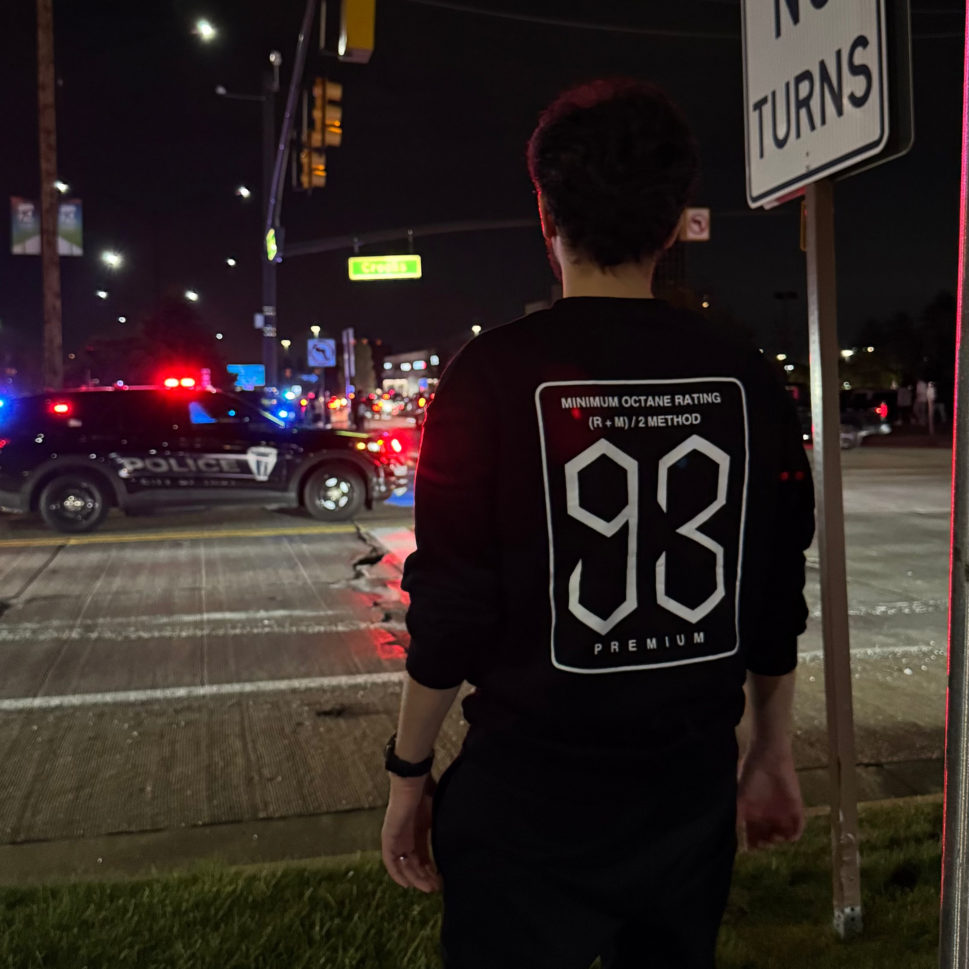 Person standing near a 'No Turns' sign at night with police cars and lights in the background.